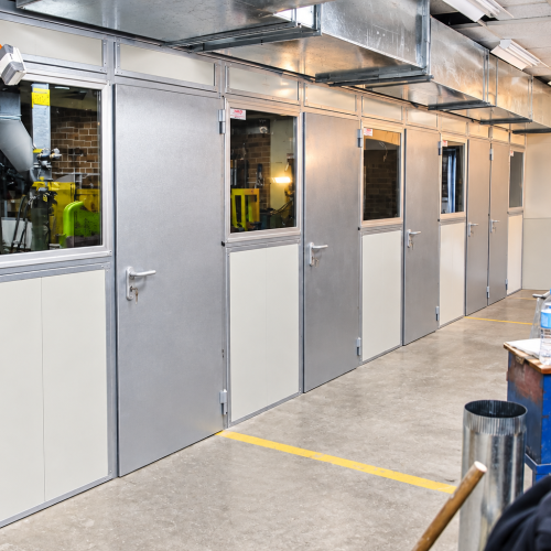 Flexshield enclosed acoustic work bays with glazed doors, modular acoustic panels and overhead ventilation ducting in a workshop.