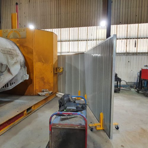 Sonic System mobile acoustic panel with perforated metal face and castor wheels positioned beside heavy industrial machinery.