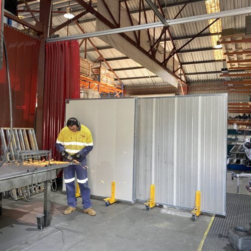 Worker using an angle grinder in a fabrication workshop with Sonic System mobile acoustic panels positioned behind the work area.