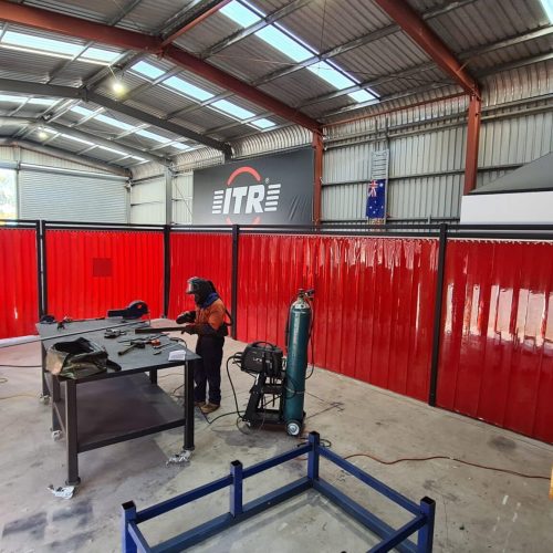 Red Weldflex™ Strip Curtains surrounding a welding workstation inside the ITR Pacific workshop.