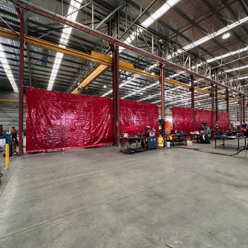 Red Weldflex™ sheet curtains dividing multiple work areas inside a large industrial fabrication workshop.