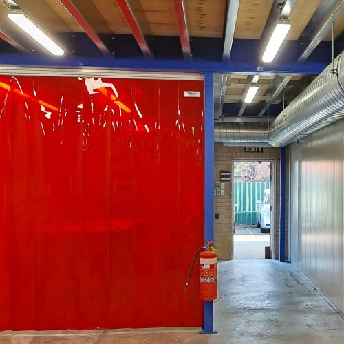 Red Weldflex™ sheet curtains hanging from overhead tracks to separate work areas inside an industrial facility.